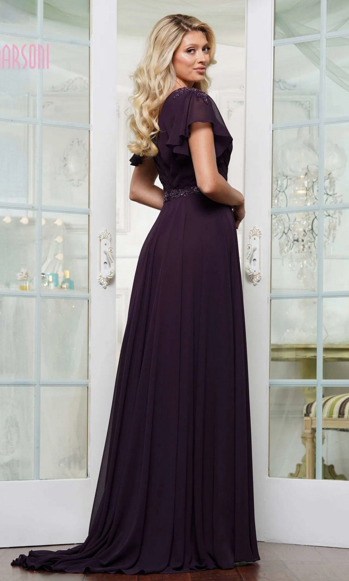 A woman stands indoors wearing the Marsoni M328, a long chiffon formal dress with an a-line skirt and beaded waistband, looking over her shoulder at the camera.